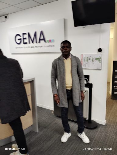 Un homme debout devant un bureau avec un panneau GEMA, portant un pull et des chaussures blanches.